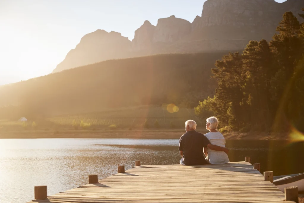 Elderly couple sitting together on a dock, overlooking a serene lake with mountains in the background during sunset.